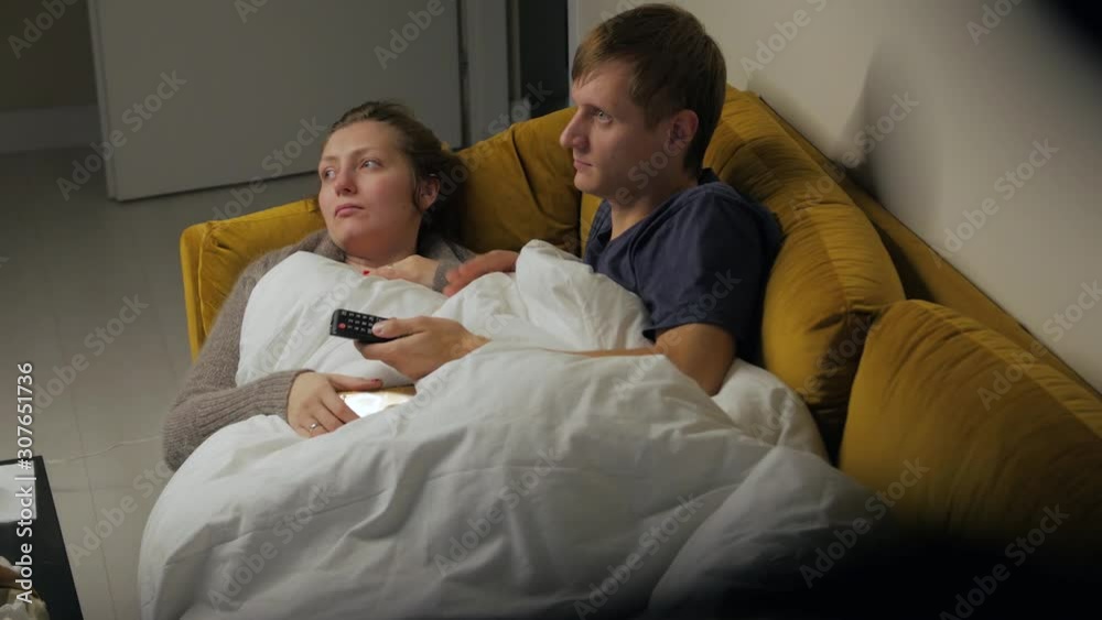 Couple watching TV on sofa talking lay on yellow couch young woman and man covered white blanket middle shot Couple watching TV on sofa talking lay on yellow couch young woman and man covered white blanket middle shot