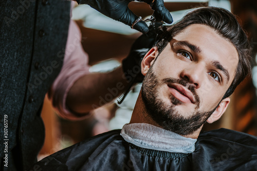 selective focus of barber in latex gloves holding scissors while styling hair of man