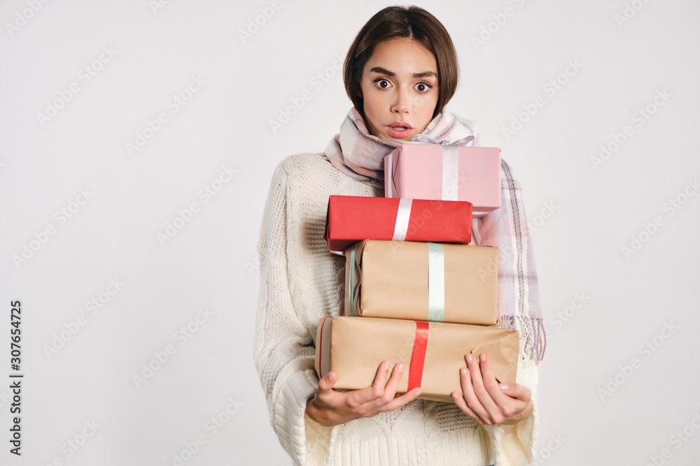 Attractive shocked girl in cozy sweater with scarf holding gift boxes amazedly looking in camera over white background Attractive shocked girl in cozy sweater with scarf holding gift boxes amazedly looking in camera over white background