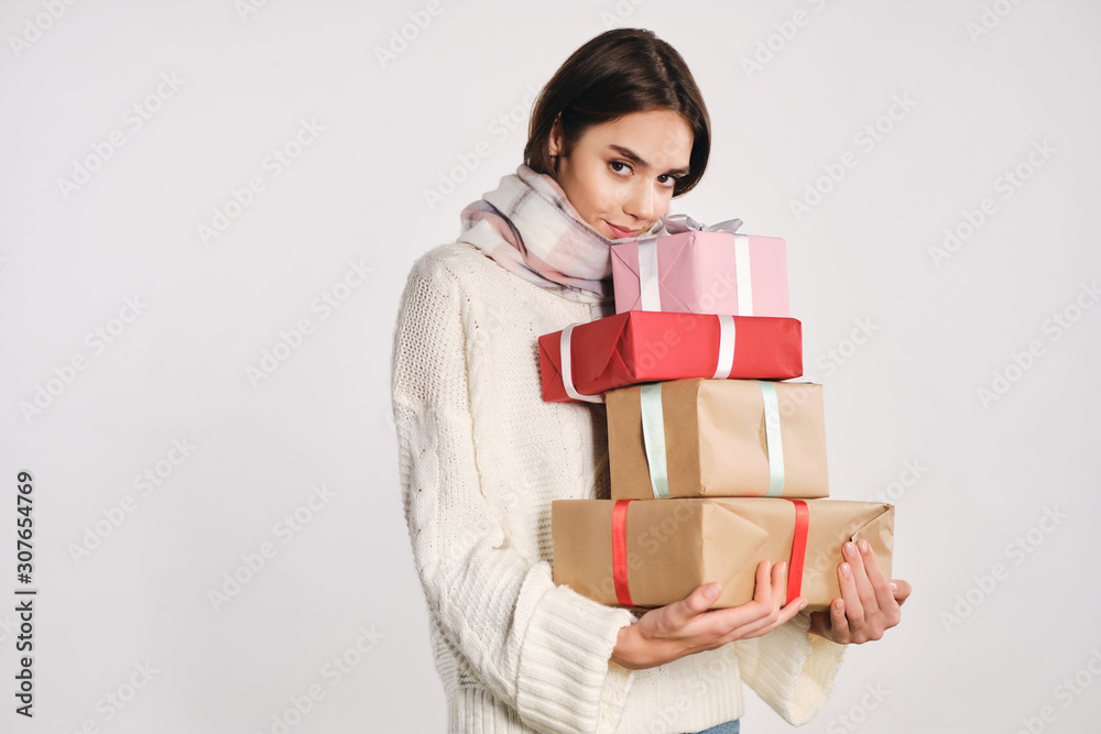 Beautiful cute girl in sweater with scarf holding gift boxes dreamily looking in camera over white background Beautiful cute girl in sweater with scarf holding gift boxes dreamily looking in camera over white background