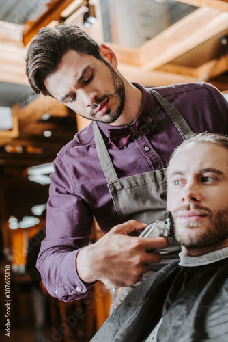 handsome barber holding trimmer while shaving bearded man