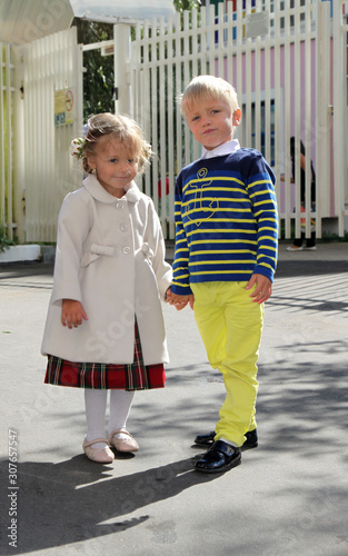 Adorable little girl in coat holds boy hand near kindergarten on sunny day. Childhood friendship