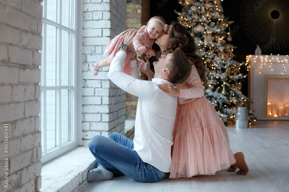 A happy family, father, mother and little daughter, spend time together on Christmas evening in a bright room indoors against a background of a beautifully decorated Christmas tree with garlands and g