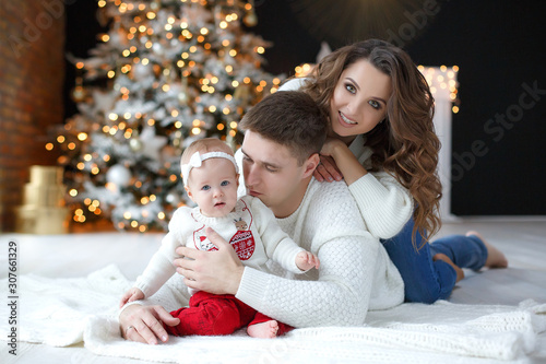 A happy family, father, mother and little daughter, spend time together on Christmas evening in a bright room indoors against a background of a beautifully decorated Christmas tree with garlands and g