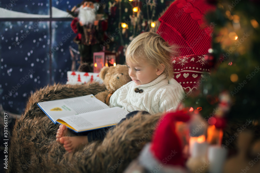 Cute toddler boy, sitting around christmas tree in cozy chair, reading book Cute toddler boy, sitting around christmas tree in cozy chair, reading book