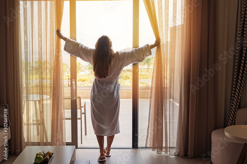 Young woman wearing white bathrobe opening curtains in luxury hotel room