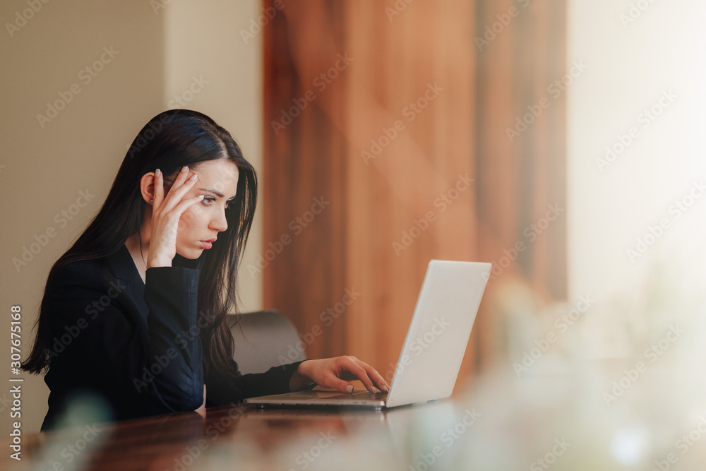 Young attractive emotional girl in business-style clothes sitting at a desk on a laptop and phone in the office or auditorium Young attractive emotional girl in business-style clothes sitting at a desk on a laptop and phone in the office or auditorium