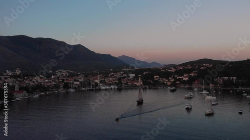 Aerial view of a small fishing boat sailing through Cavtat harbor, calm water at sunset with views of the Mountainous landscape and towns coastline.