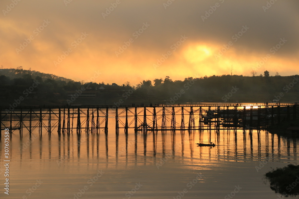 A little boat and wooden bridge with mountain landscape over the river at Sunrise in Thailand.