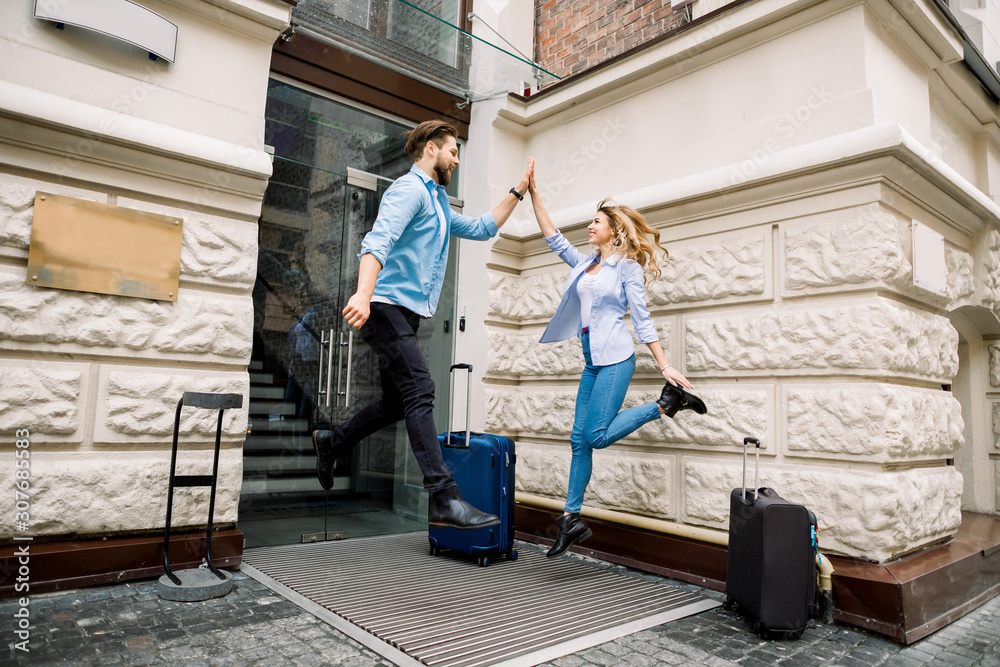 Young cheerful couple with suitcases jumping and giving five, having fun, before arriving to the hotel. Couple having fun outdoors, near the old city building Young cheerful couple with suitcases jumping and giving five, having fun, before arriving to the hotel. Couple having fun outdoors, near the old city building