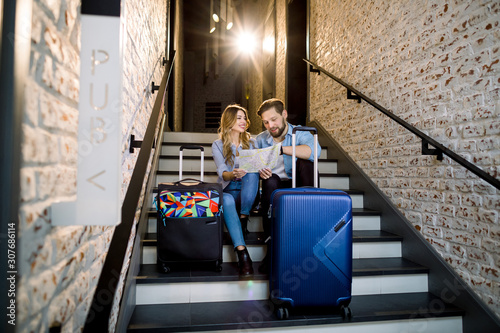 attractive smiling couple of man and woman in smart casual business style with two suitcases, sitting on stairs in stylish loft hotel hall indoors, looking together on city map © sofiko14