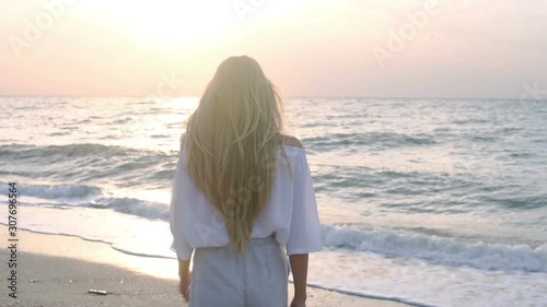 Back view of beautiful slim blonde with long hair in a white blouse walkng along the coast at sunset photo