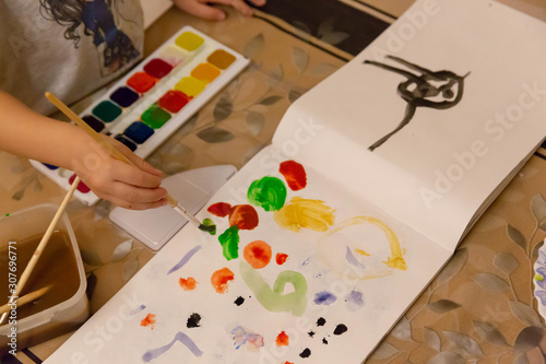 Children's hand with a brush with a wooden handle draws on a white sheet of paper album for drawing, lying on the table, colorful watercolor paints in children's art school.