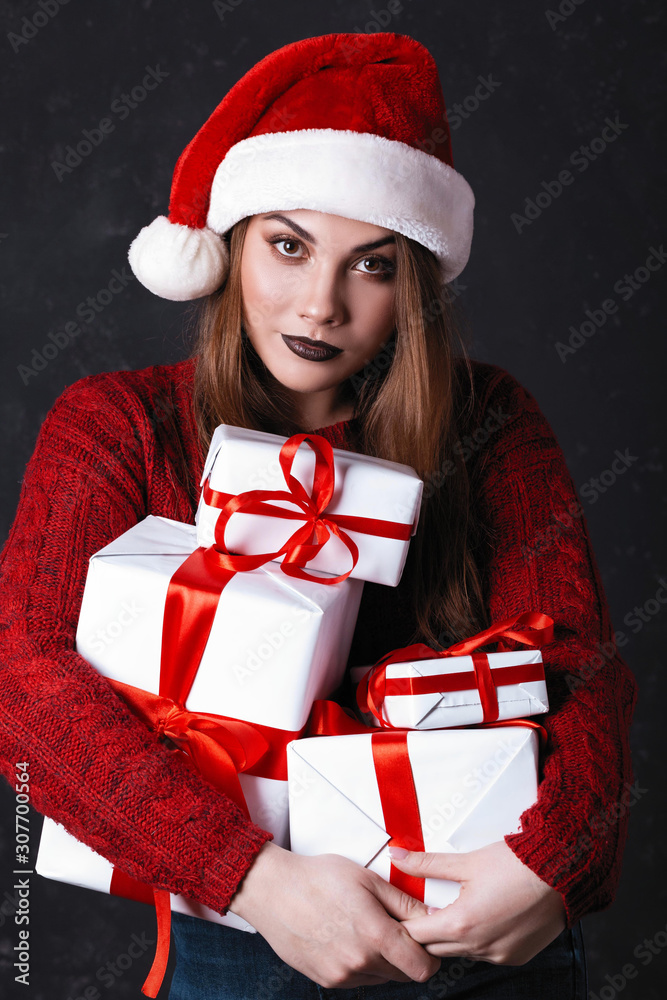 Lovely girl with make-up in a burgundy sweater and santa claus hat. Posing on a dark background holds gifts on a Christmas tree, the concept of Christmas and New Year Lovely girl with make-up in a burgundy sweater and santa claus hat. Posing on a dark background holds gifts on a Christmas tree, the concept of Christmas and New Year