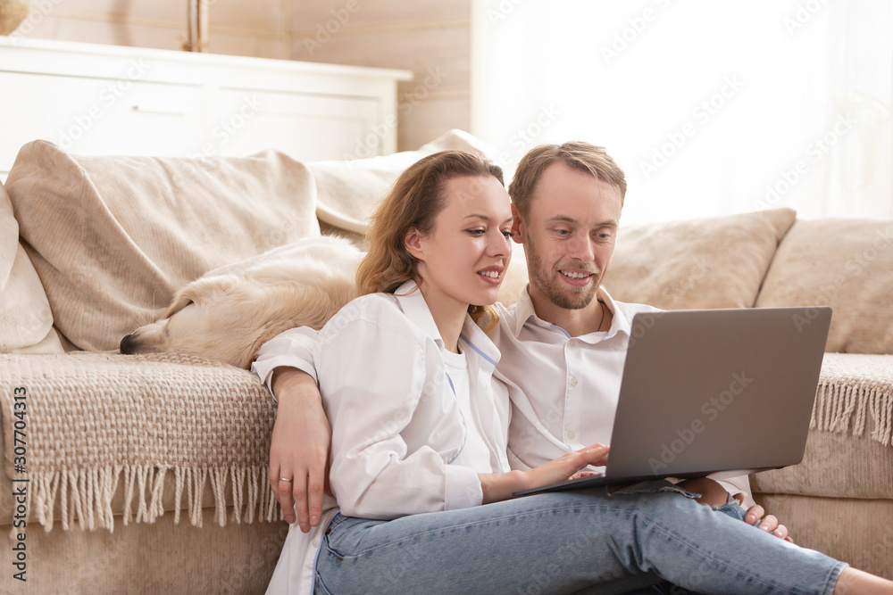 Young happy positive married couple charming woman and young man all sit on the floor next to their beloved dog and watch cheap airline tickets for summer holidays on the Internet using a laptop