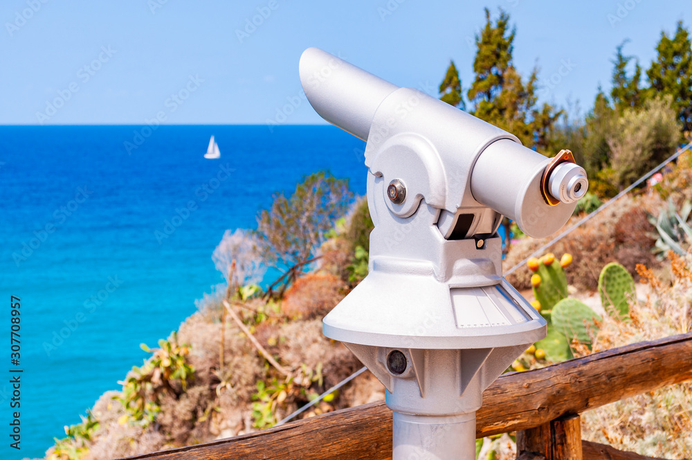 Stationary touristic telescope made of metal on one leg standing on the edge of a cliff near the wooden logs fence and observes the Tyrrhenian sea with floating white sailing yacht in Tropea