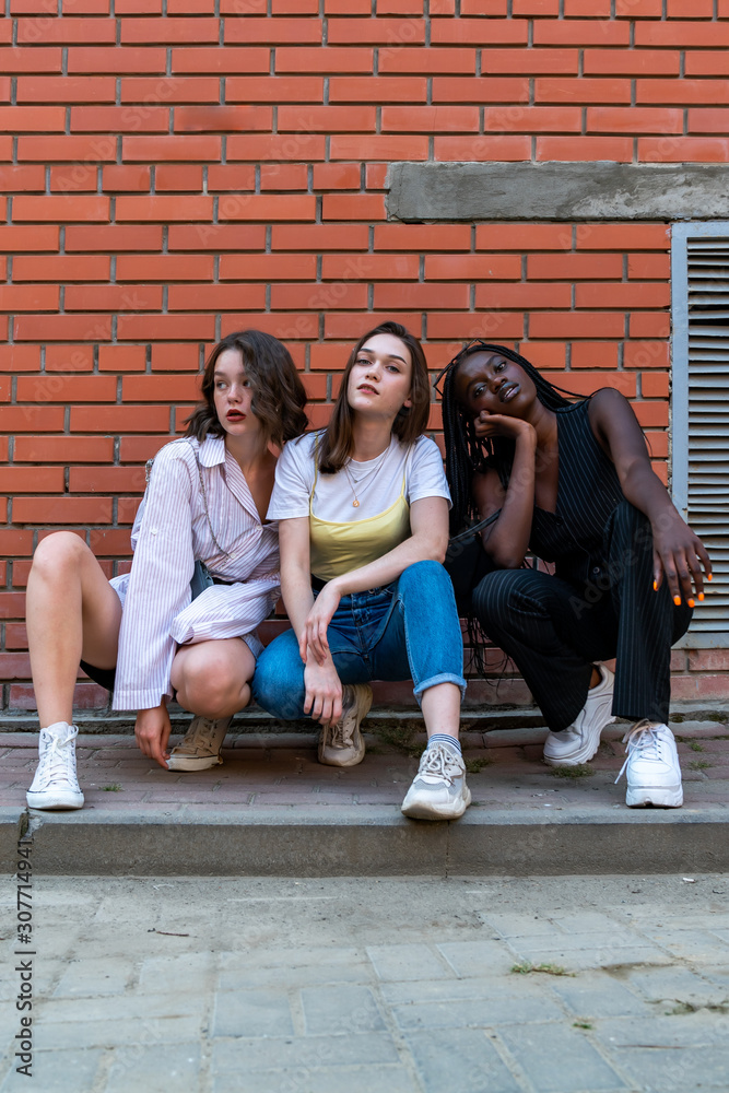 Multi ethnic stylish portrait of young girls in casual clothes that crouched and looking at the camera isolaed over red brick wall