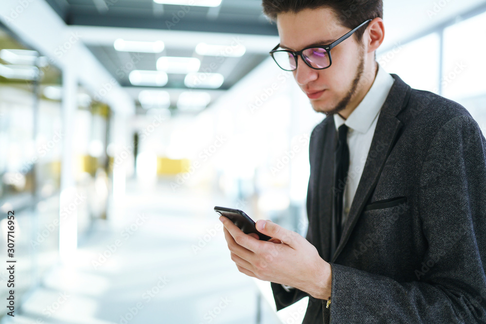 Man's hands using mobile phone standing in the modern office. Businessman holds telephone in hand. Stylish man in a suit with phone in hands prints a message on the phone. 