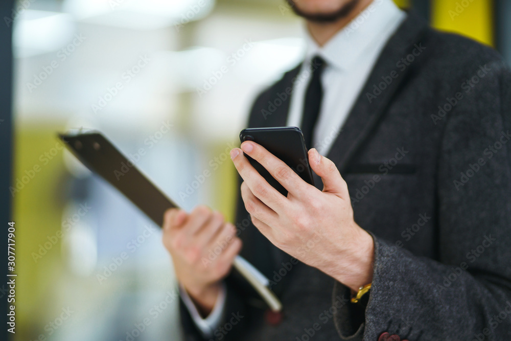 Man's hands using mobile phone standing in the modern office. Businessman holds telephone in hand. Stylish man in a suit with phone in hands prints a message on the phone. Man's hands using mobile phone standing in the modern office. Businessman holds telephone in hand. Stylish man in a suit with phone in hands prints a message on the phone.