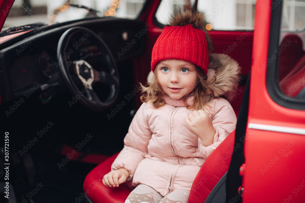 Smiling cute winter girl in red hat sitting in car having fun medium shot. Happy beautiful female baby in warm clothing having positive emotion outdoor surrounded by snowflakes enjoying childhood