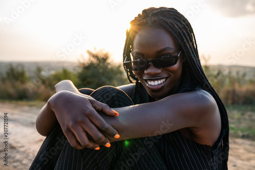 Close up portrait of the young happy african girl in sunglasses that sits on the dirt road, smiling and looking at the camera