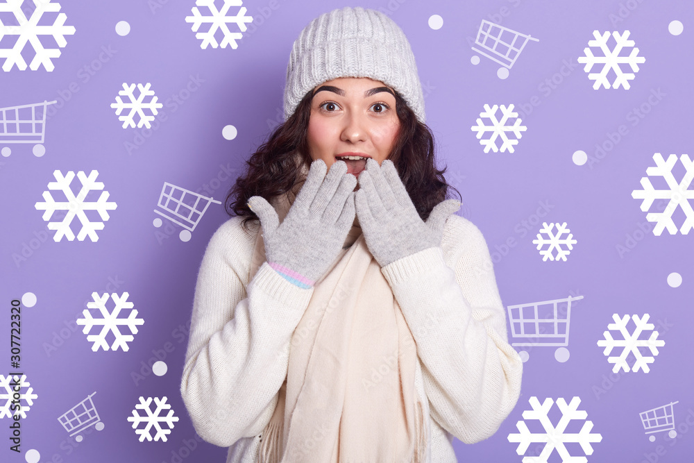 Close up portrait of bruunetee girl wearing warm winter clothes, knitted sweater, hat and mittens, having astonished facial expression, attractive female keeping hands on opened mouth, looks surprised Close up portrait of bruunetee girl wearing warm winter clothes, knitted sweater, hat and mittens, having astonished facial expression, attractive female keeping hands on opened mouth, looks surprised
