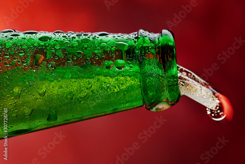 Close-up of the neck of a green bottle in drops and a splash of beer in the shape of a tongue on a red background