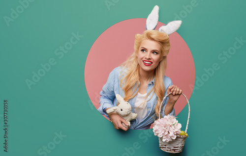 Young pin-up woman with easter bunny ears with a basket of colored eggs crawls out through a round hole in the wall. Menthol or turquoise color background. Holiday card blank