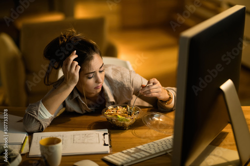 Billede på lærred Young woman feeling displeased while eating salad and working late at home