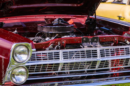 close up view of red classic muscle car front with open hood, headlight, Radiator, Chrome Bumper, big round air intake filter, and other engine parts