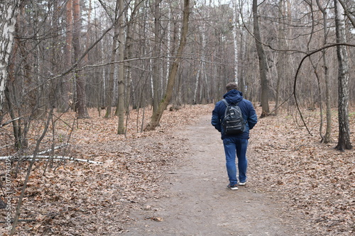 A man in blue clothes with a black backpack leaves on the beaten road in the gray autumn forest.