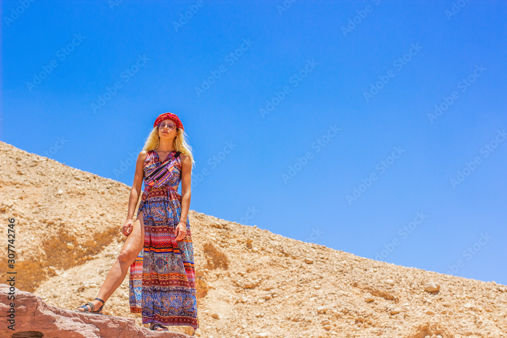 sexual European white woman in long fashion colorful dress looking on you from above in desert environment on blue sky background with empty copy space for your text 
