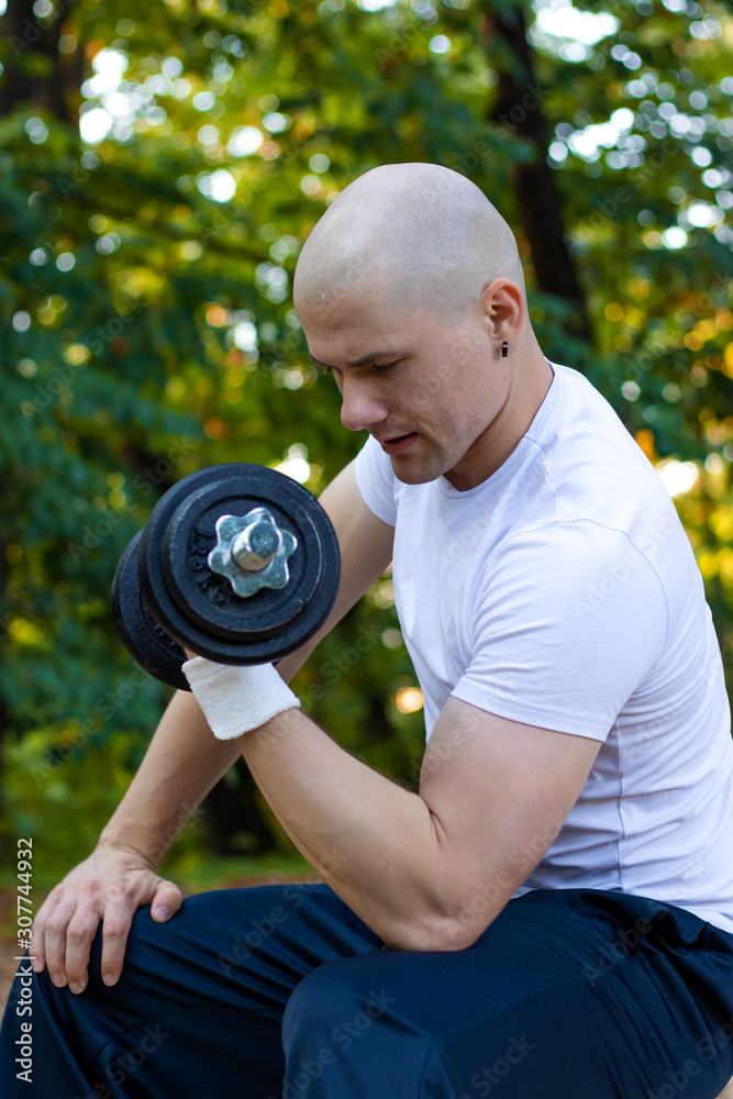 Handsome muscular young athlete man working out, weight training with dumbbell in city park. Fitness, sport, training and lifestyle concept.
