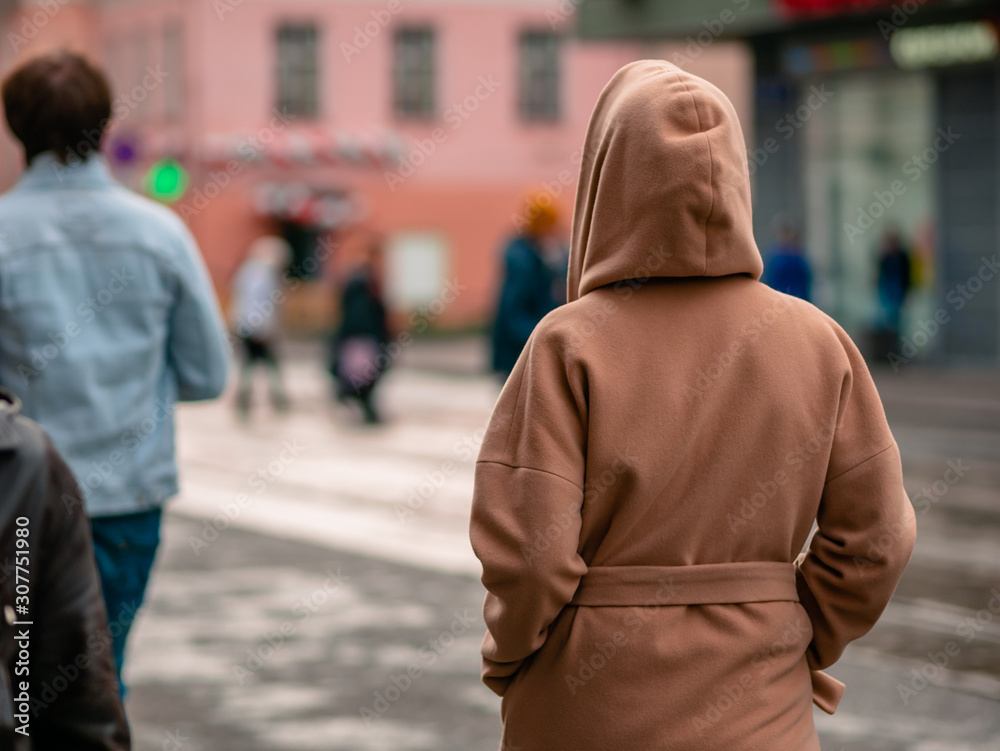 A woman walks down the street in a demi-season brown warm coat with a hood. Unrecognizable girl walks on the road. The view from the back. Warm autumn day. Blurry silhouettes of passers-by A woman walks down the street in a demi-season brown warm coat with a hood. Unrecognizable girl walks on the road. The view from the back. Warm autumn day. Blurry silhouettes of passers-by