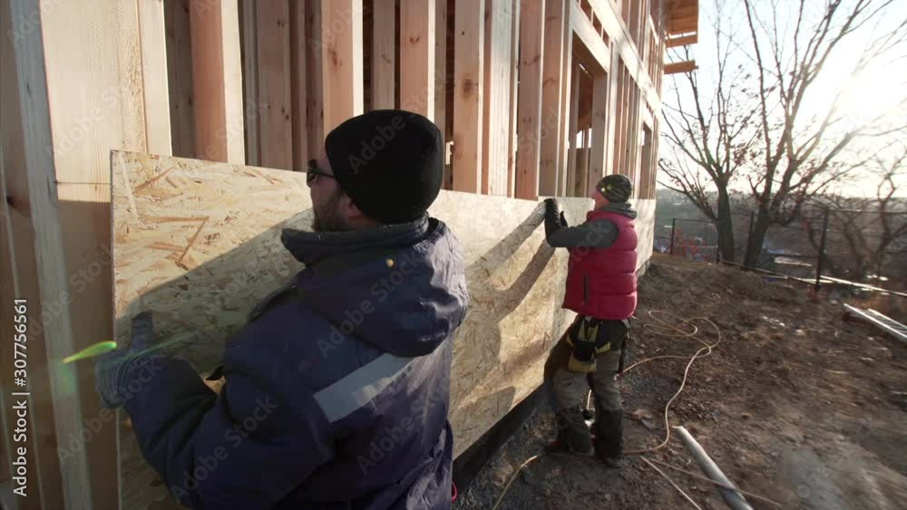 Two workers are covering the wooden house frame with plywood. Wooden frame residential house under construction