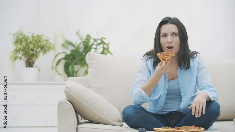 Hungry woman is switching on the TV program, while sitting on a sofa. She is eating pizza. 4K. Hungry woman is switching on the TV program, while sitting on a sofa. She is eating pizza. 4K.