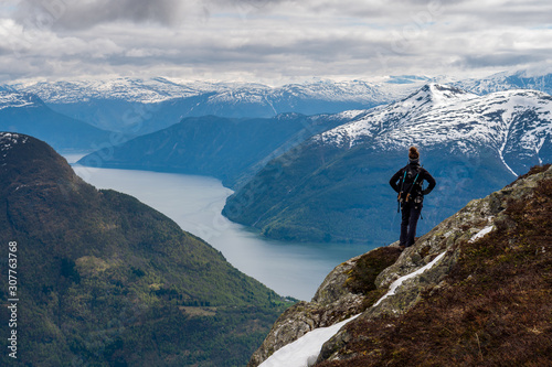 An explorer stands atop a mountain looking across a sweeping view of Norwegian fjords and snow capped mountains