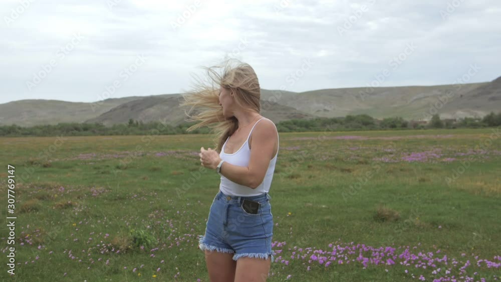 Camera follow hipster young woman in white t-shirt short jeans fluttering long hair. One girl dancing on field of grass portable music speaker smartphone. Happy drunk from life, youth and happiness.
