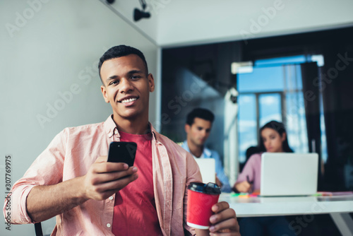 Portrait of cheerful male financial expert with tasty caffeine beverage and digital smartphone smiling at camera, happy millennial man trainee using office internet connection on mobile gadget