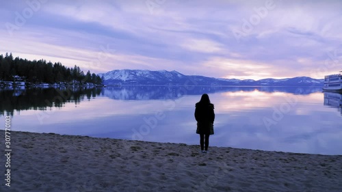 Aerial: Woman standing on a lake foreshore looking across at snowy mountains. Lake Tahoe, Californina, USA. 1 April 2019 photo