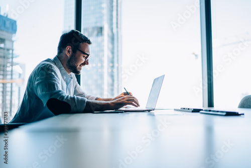 Side view of successful office worker sitting at table desktop in office company interior and using modern netbook for searching trade information, happy Caucasian male investor checking website news