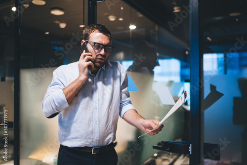 Financial marketer holding paper documents in hand and making smartphone conversation with executive employee for discussing mistakes in report, Caucasian office worker analyzing capital investment