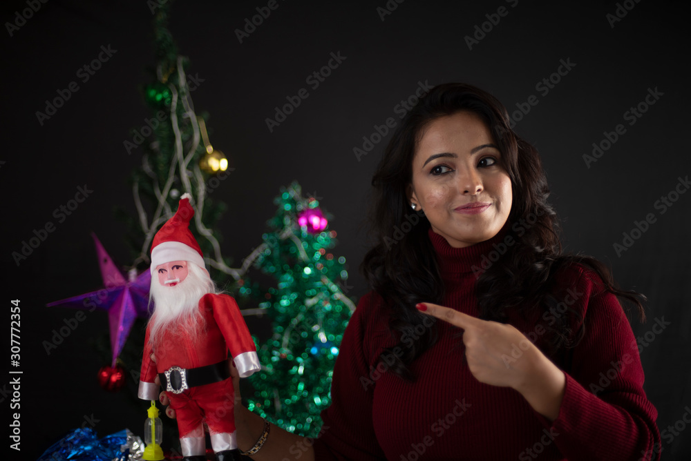 Young and attractive brunette Indian Bengali woman in western woolen highneck maroon sweater showing toy Santa clause in decorative Christmas tree background.Indian lifestyle and Christmas celebration Young and attractive brunette Indian Bengali woman in western woolen highneck maroon sweater showing toy Santa clause in decorative Christmas tree background.Indian lifestyle and Christmas celebration