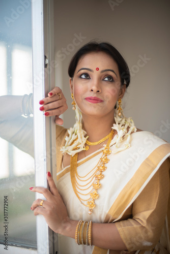 An young and attractive Indian woman in white traditional wear is smiling while standing in front of a glass window for the celebration of Onam/Pongal in white background. Indian lifestyle. © abir