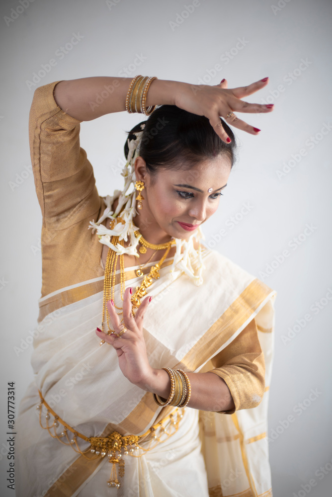 Obraz premium An young and attractive Indian woman in white traditional wear dancing traditional dance form for the celebration of Onam/Pongal in white background. Indian lifestyle.