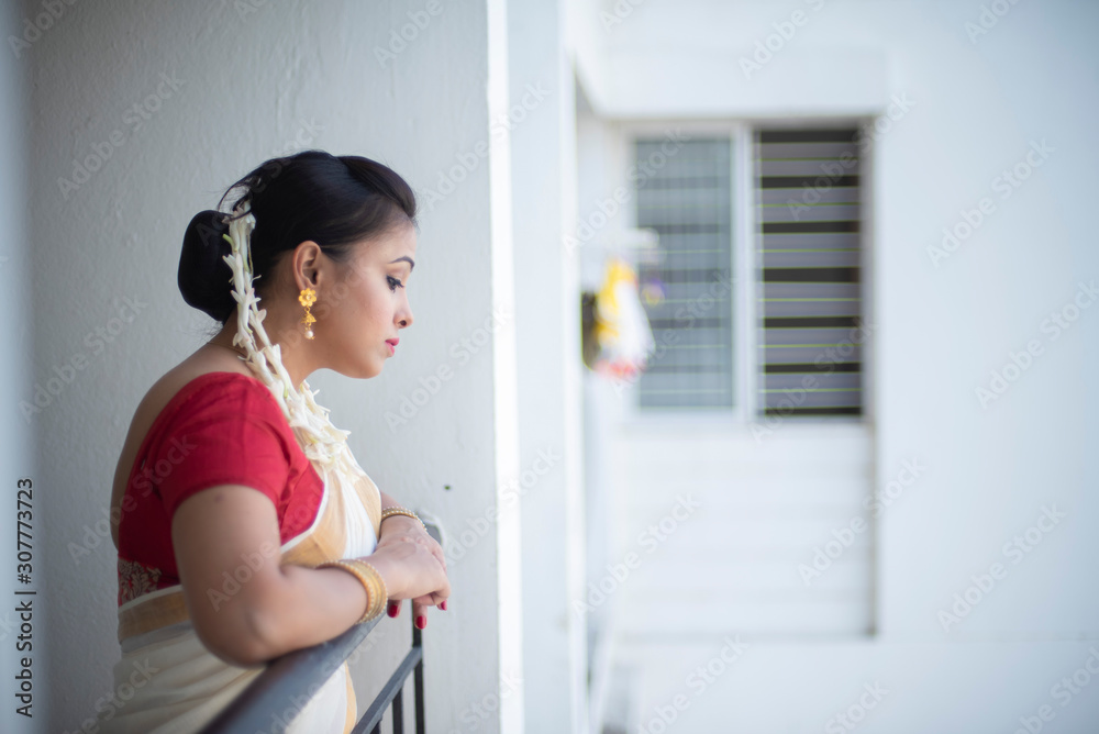 An young and attractive Indian woman in white traditional sari and red blouse and flowers is standing in a balcony for the celebration of Onam/Pongal. Indian lifestyle. An young and attractive Indian woman in white traditional sari and red blouse and flowers is standing in a balcony for the celebration of Onam/Pongal. Indian lifestyle.