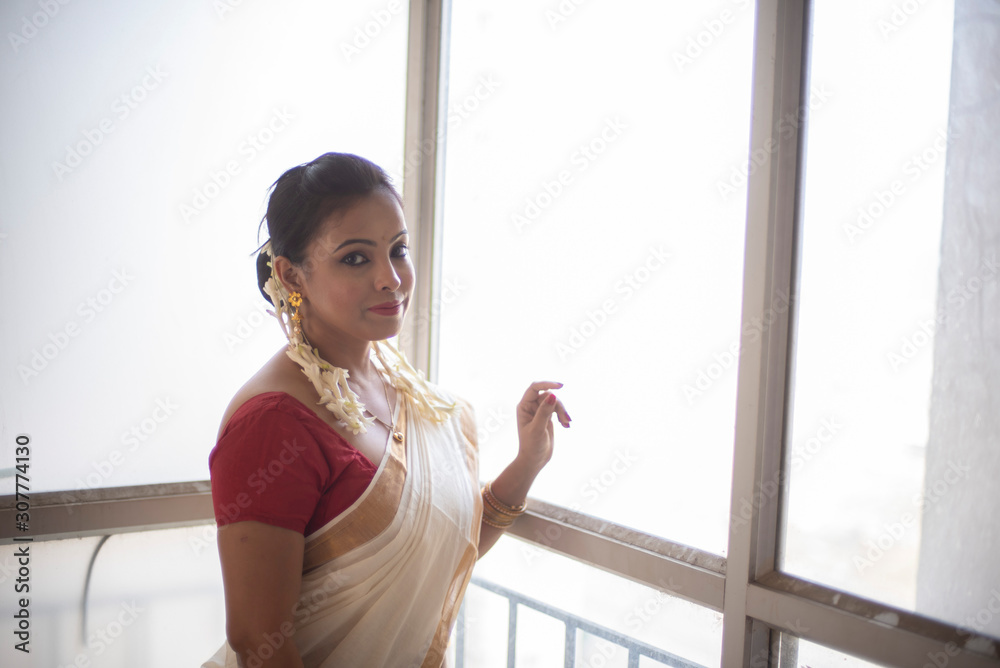 An young and attractive Indian woman in white traditional sari and red blouse and flowers standing in front of a glass window in a balcony  for the celebration of Onam/Pongal. Indian lifestyle.