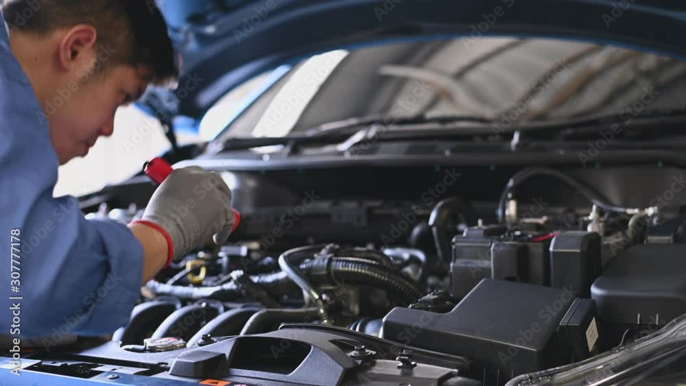Young Asian auto mechanic examining engine at car hood in auto repair workshop. Repairman checking for customer to safety maintenance support before long trip. Business and transportation concept.