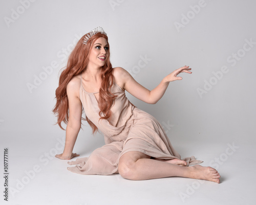full length portrait of a pretty, fairy girl wearing a nude flowy dress and crystal crown. Seated pose against a grey studio background.