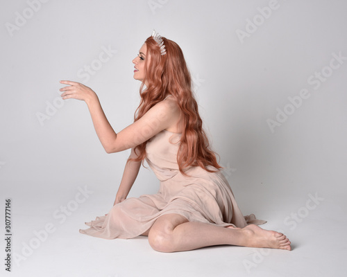 full length portrait of a pretty, fairy girl wearing a nude flowy dress and crystal crown. Seated pose against a grey studio background.